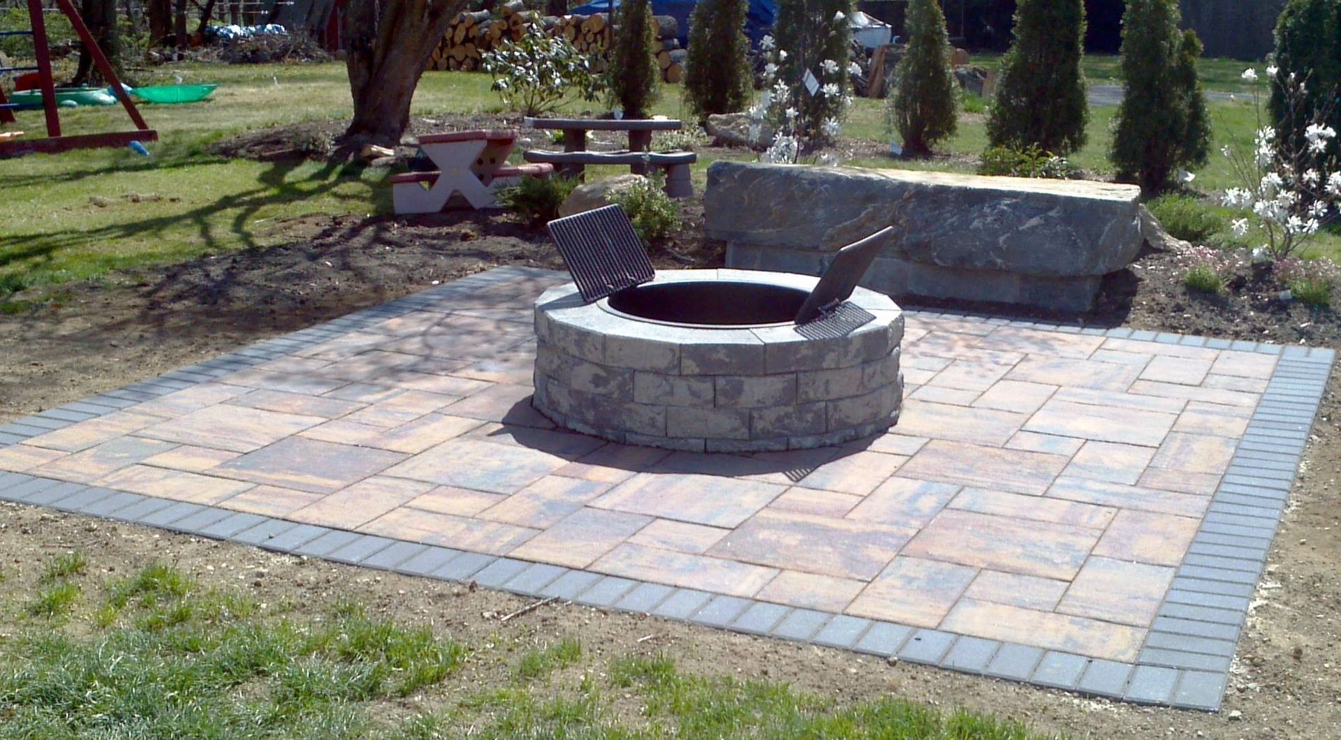 Fire pit on a paved patio with a stone bench in a backyard setting.