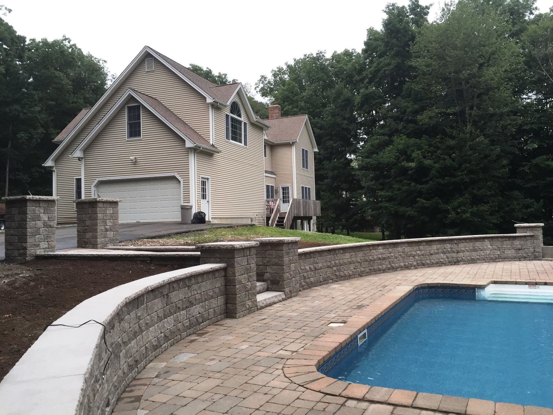 A two-story beige house with a brick pool, retaining walls, and a curved walkway.