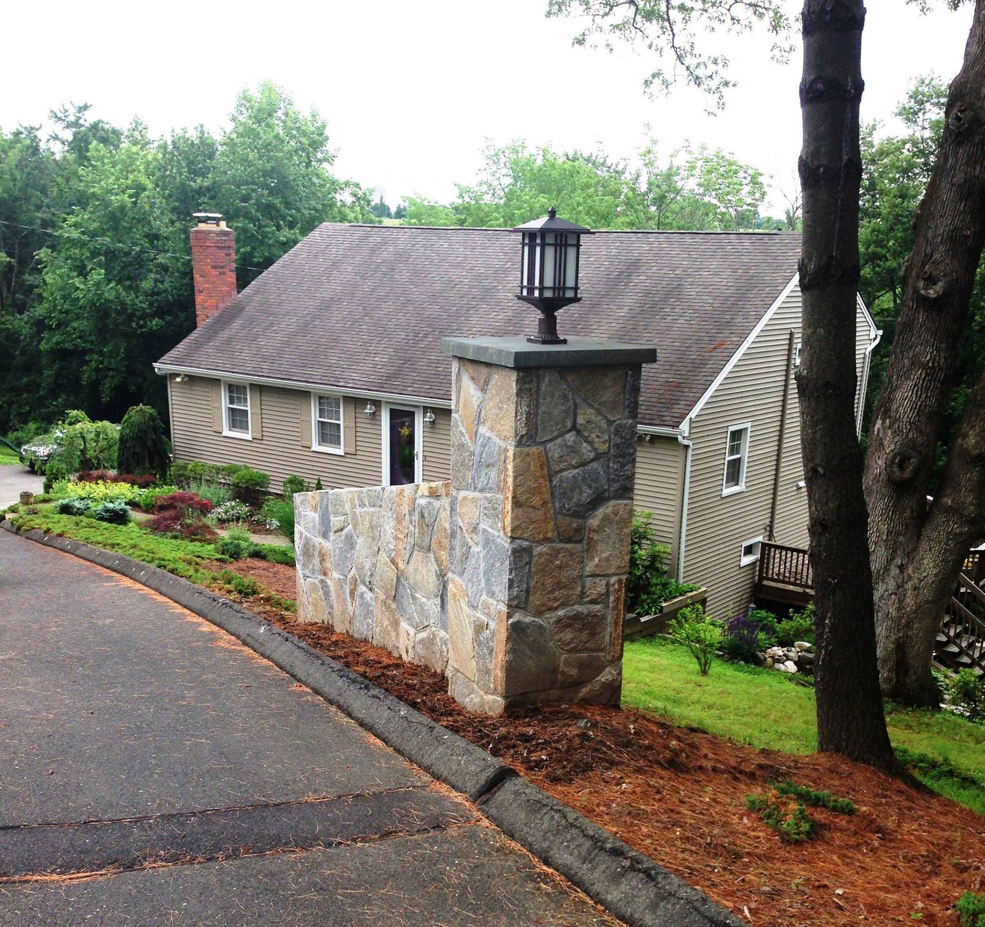 Stone pillar with lamp at a driveway entrance to a house; landscaping on both sides.