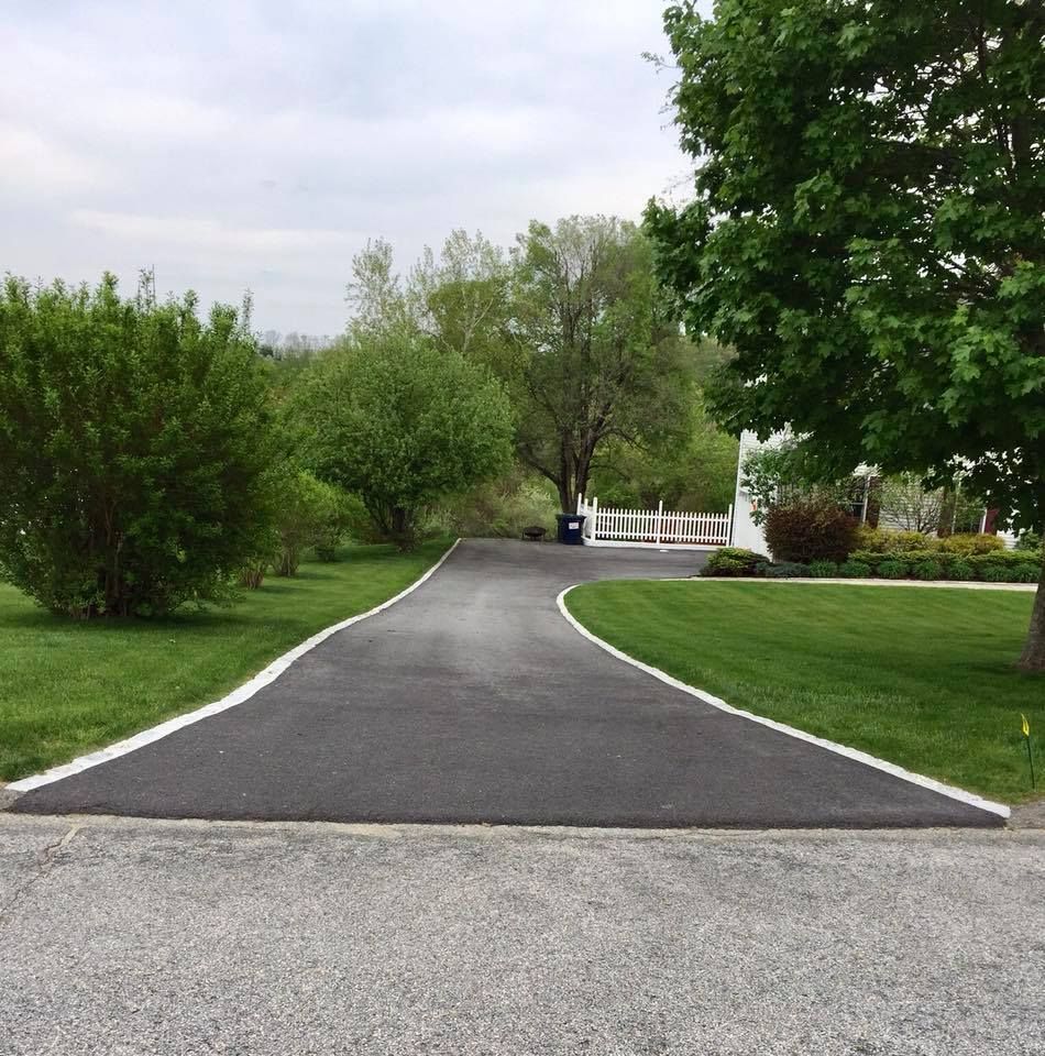 Asphalt driveway with white-lined edges curves toward a house, surrounded by green lawn and trees.