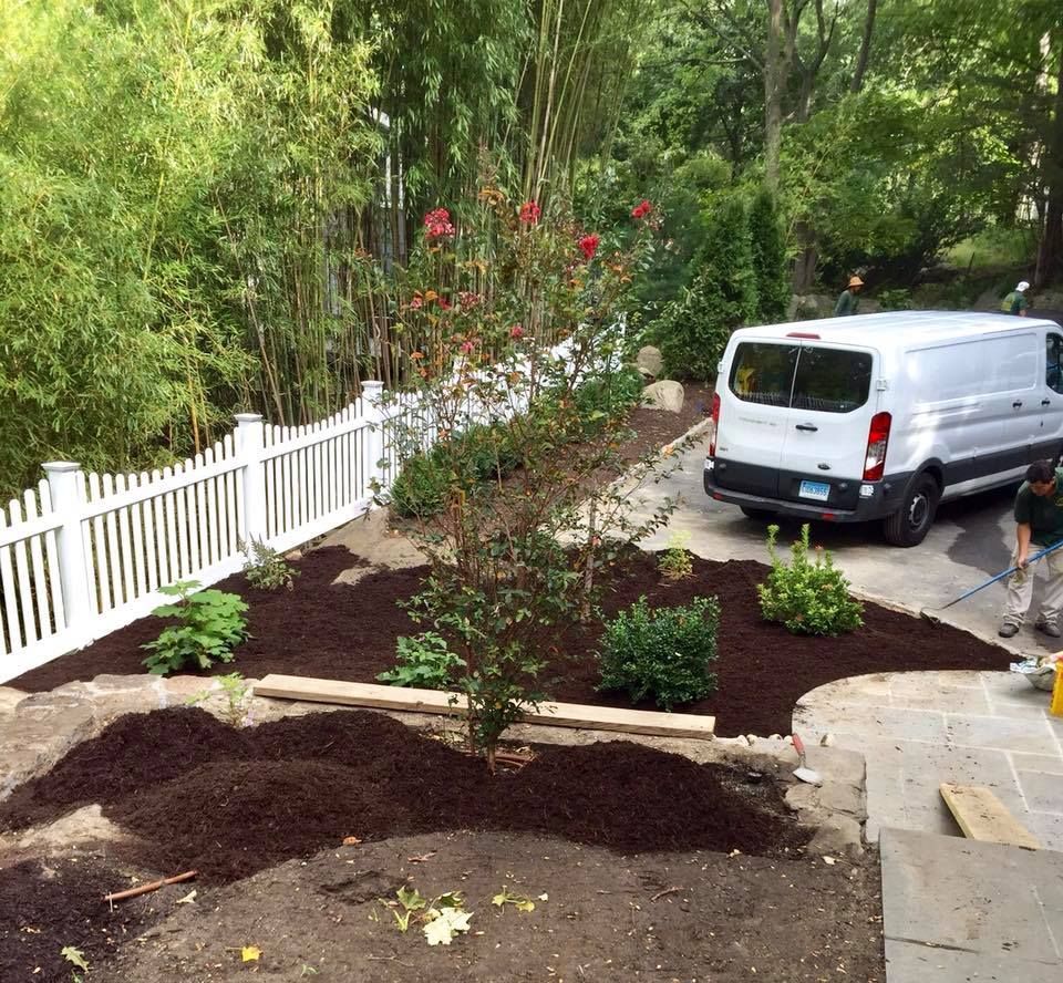 Backyard garden with newly-laid mulch and plants; white picket fence; white van parked on the driveway.