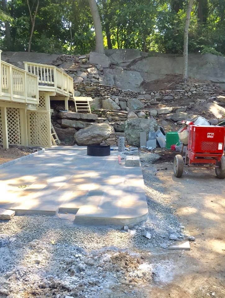 Concrete patio construction next to a wooden deck, with large rocks in the background. A red cement mixer is present.