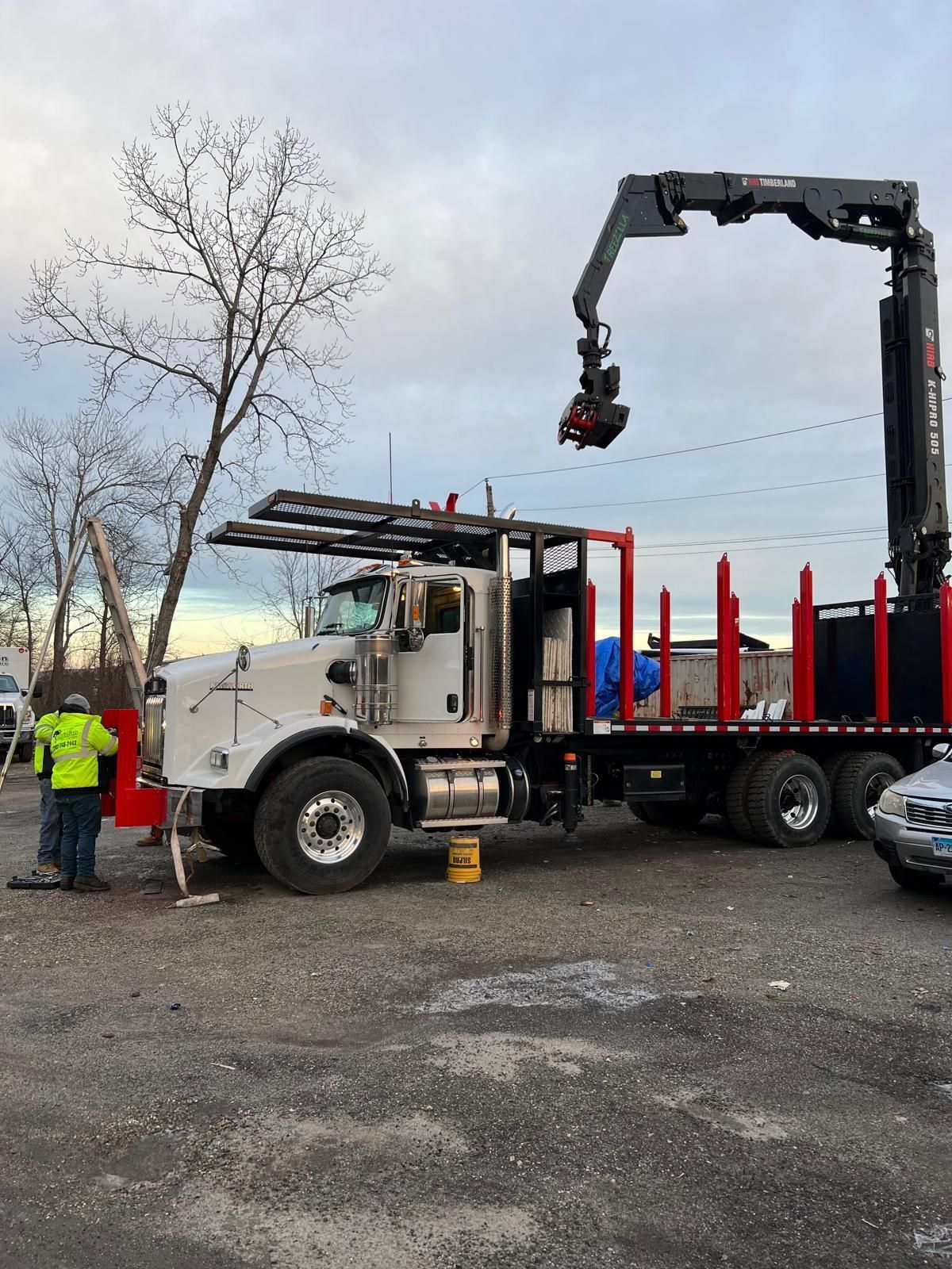 White logging truck with crane lifting logs, worker in yellow vest nearby.