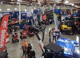 Crowded indoor showroom with tractors, farm equipment, banners, and several people browsing displays