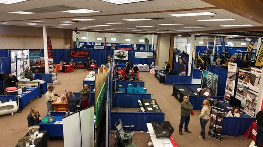 Indoor trade show booth area with tables, displays, and a few people browsing under bright ceiling lights