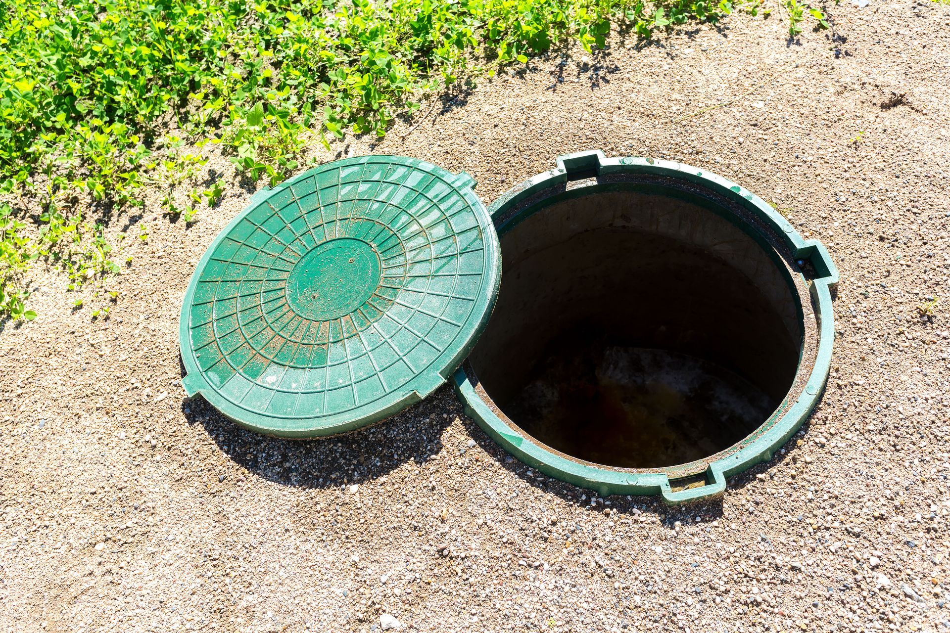 Open green manhole with lid beside it, in gravel near grass.