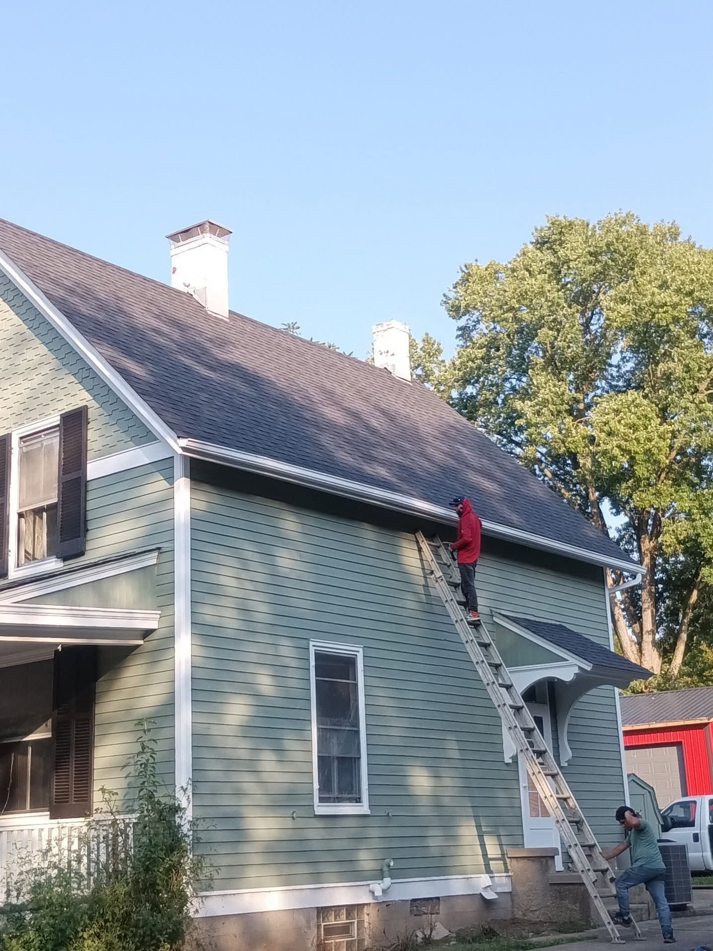 Two workers on a ladder repainting a green house exterior under a steep roof