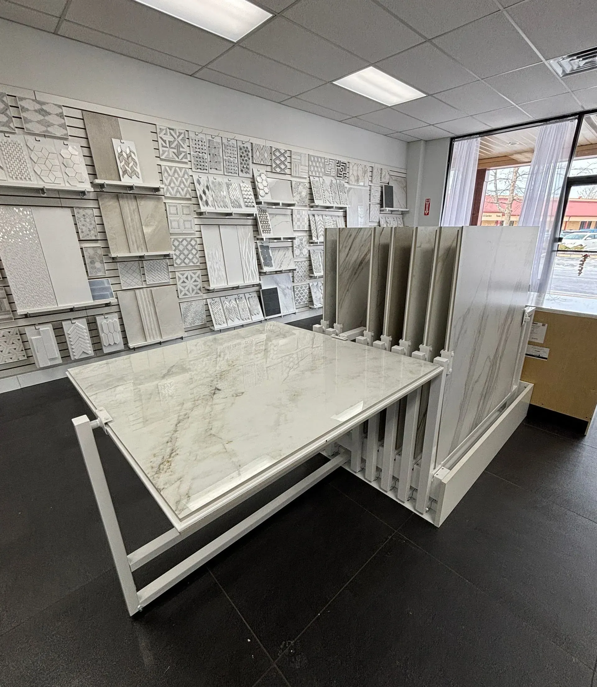 Retail store display area with white shelving, sample boards, and a table on a black carpeted floor.