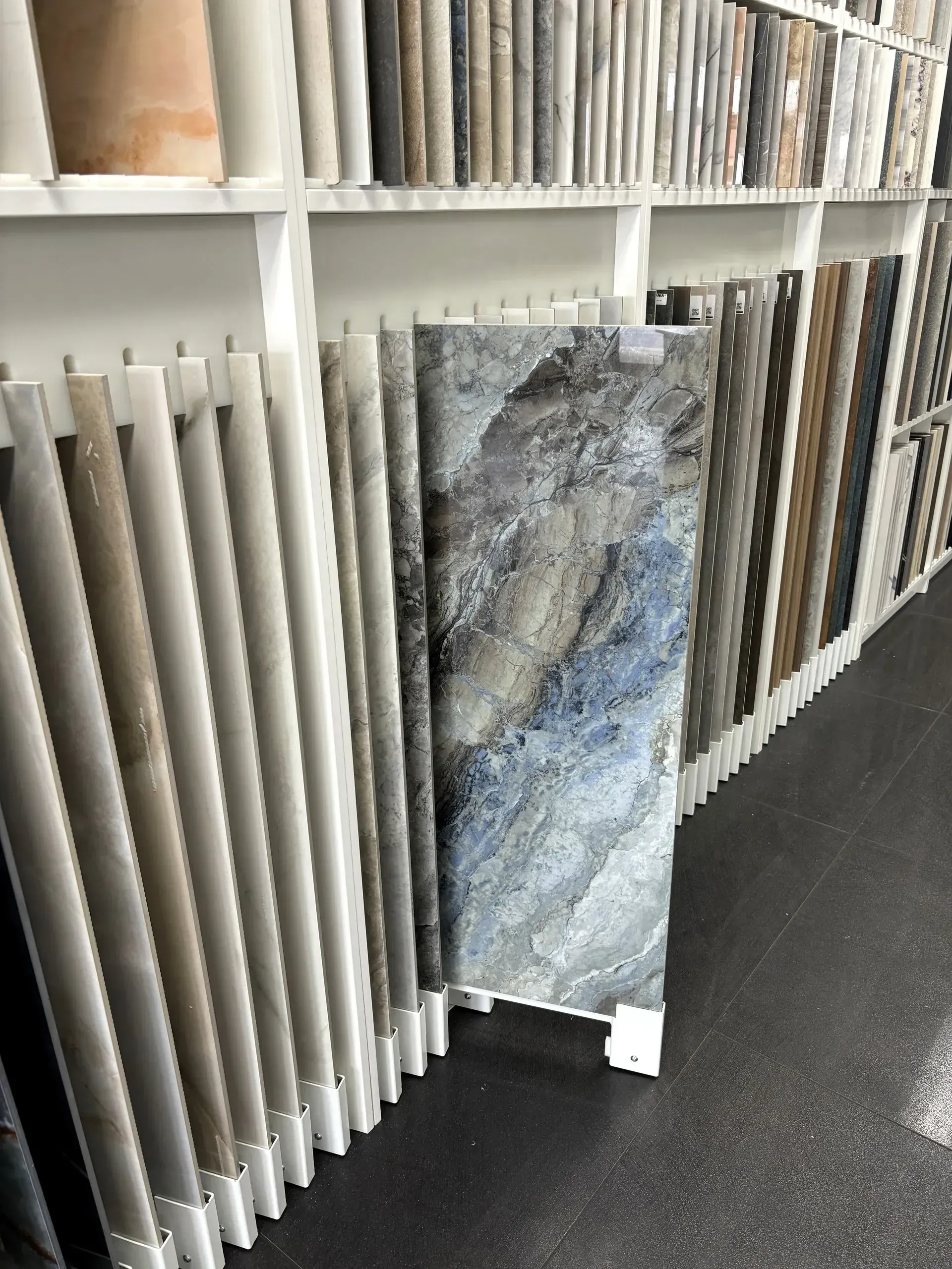 Tile showroom display with gray and beige sample slabs on white racks beside a dark floor