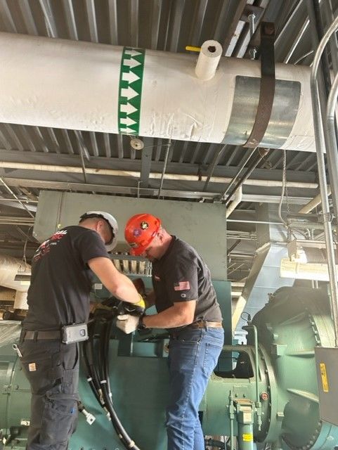 two people fixing a black pipe in a huge machine