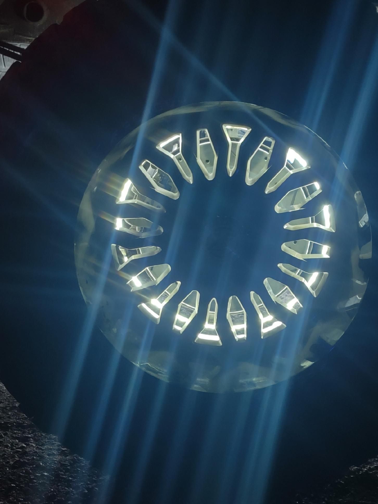 Bright light shines through a circular, patterned metal grill in a dark setting.