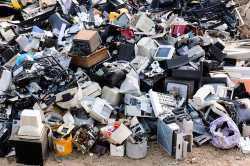 Pile of discarded electronics and scrap piled high in a junkyard, with mixed white, black, and silver appliances.