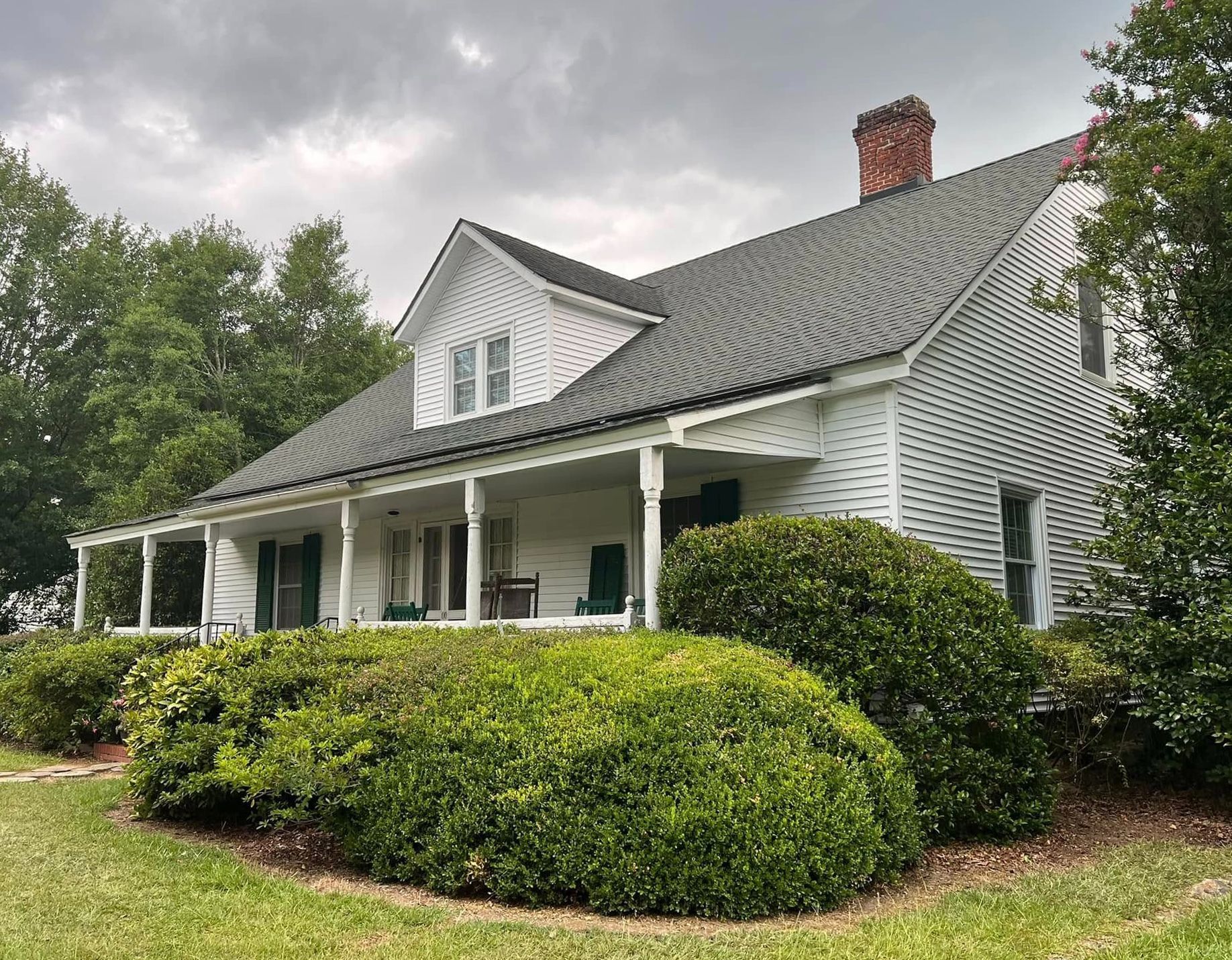 White house with a wrap-around porch, green shutters, and dark roof, surrounded by green bushes and grass.