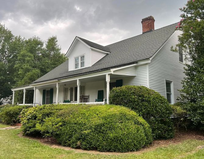 White house with a wrap-around porch, green shutters, and dark roof, surrounded by green bushes and grass.