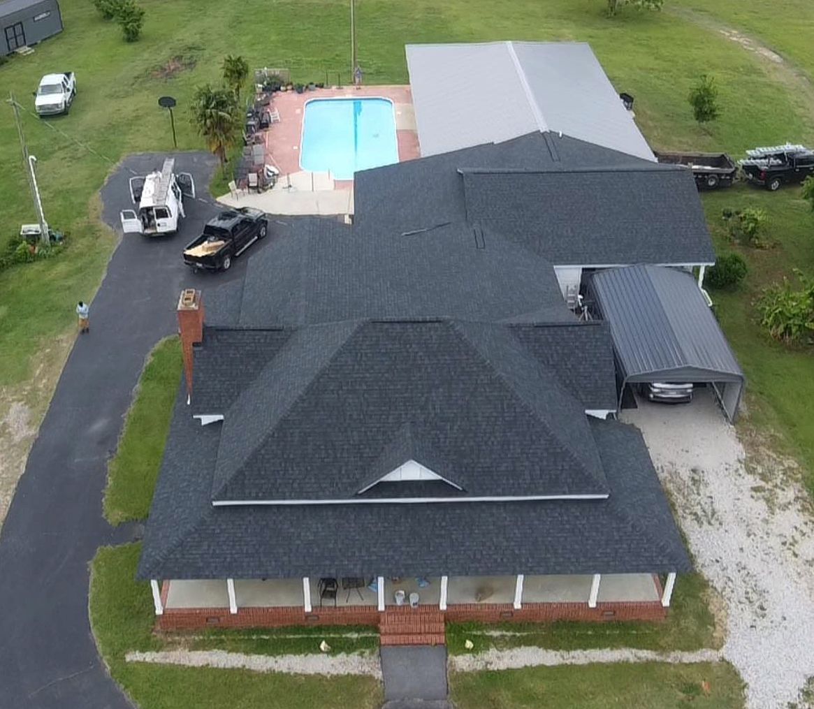 Aerial view of a house with a dark roof, driveway, swimming pool, and a car parked.