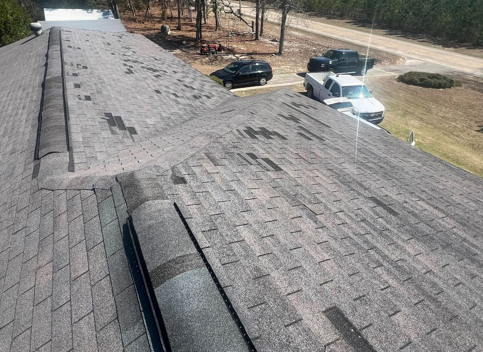 Damaged asphalt shingle roof with missing granules and vehicles parked below.