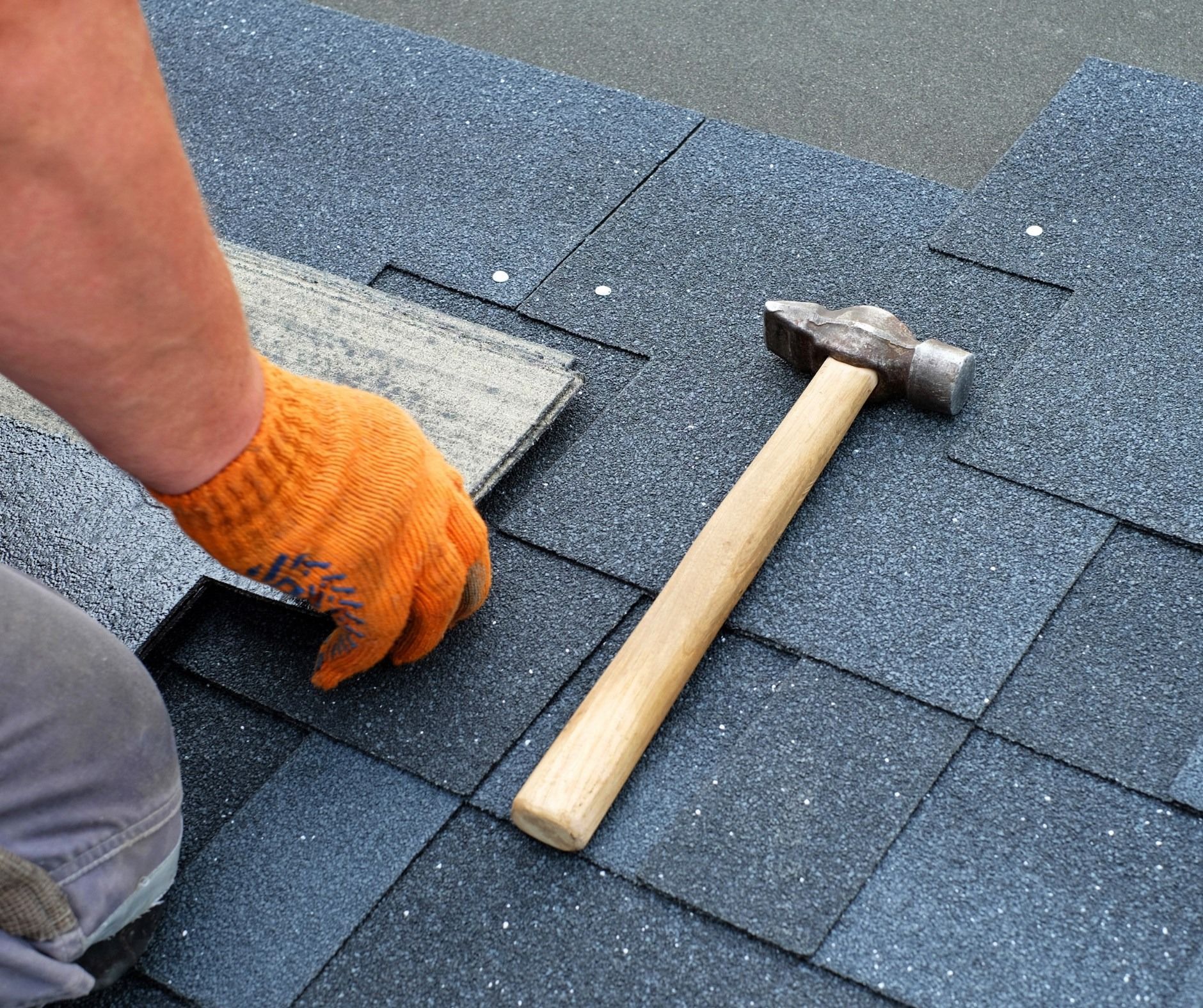 A worker in orange gloves installing dark blue shingles on a roof with a hammer.