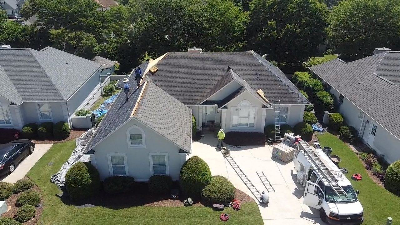 Workers replacing a roof on a light blue house. A white truck and ladder are parked in the driveway.