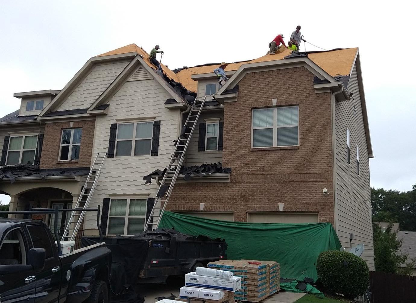 Roofers replacing shingles on a two-story house with brick and siding. Ladders, truck, and supplies are present.