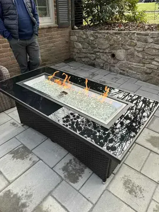 Outdoor rectangular fire table with flames, on a patio beside a brick house.