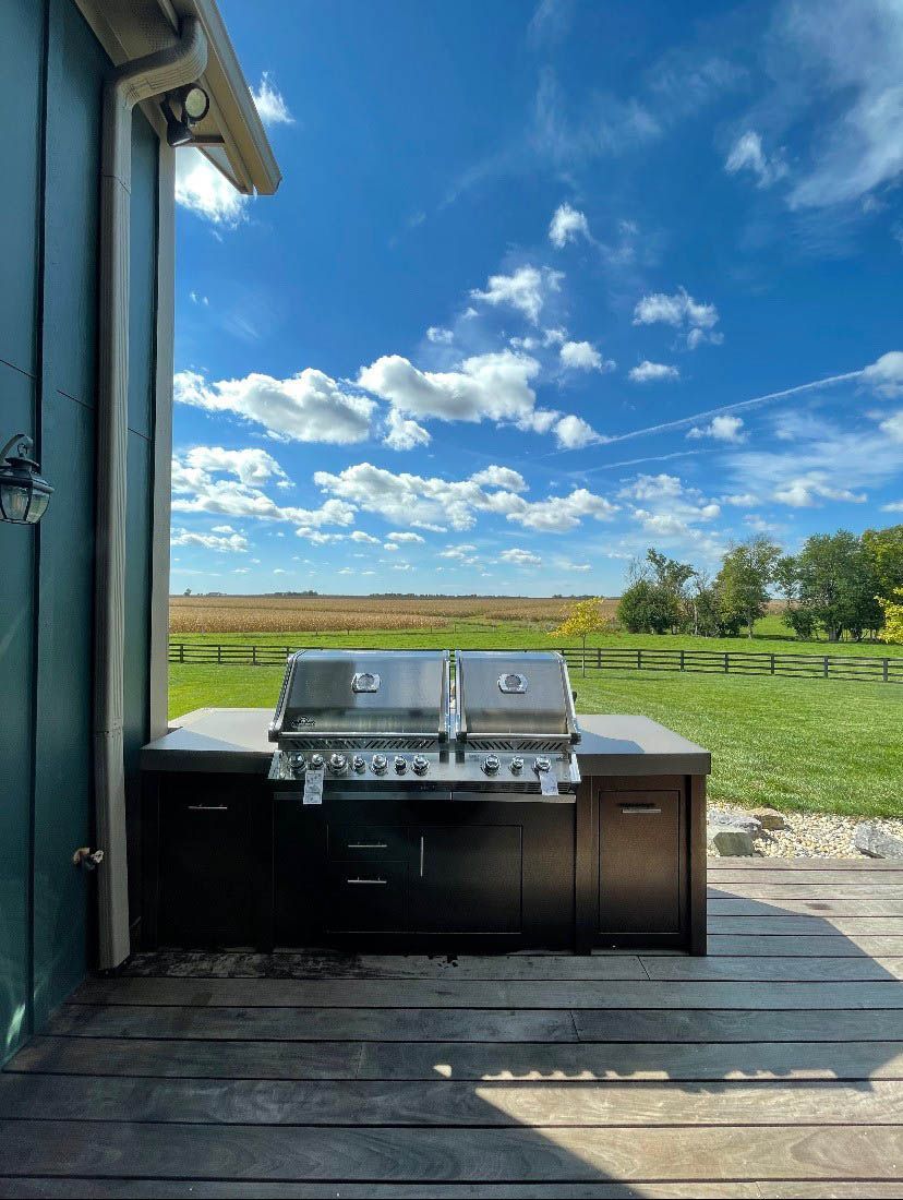 Outdoor deck with a stainless steel grill overlooking a green field and blue sky.