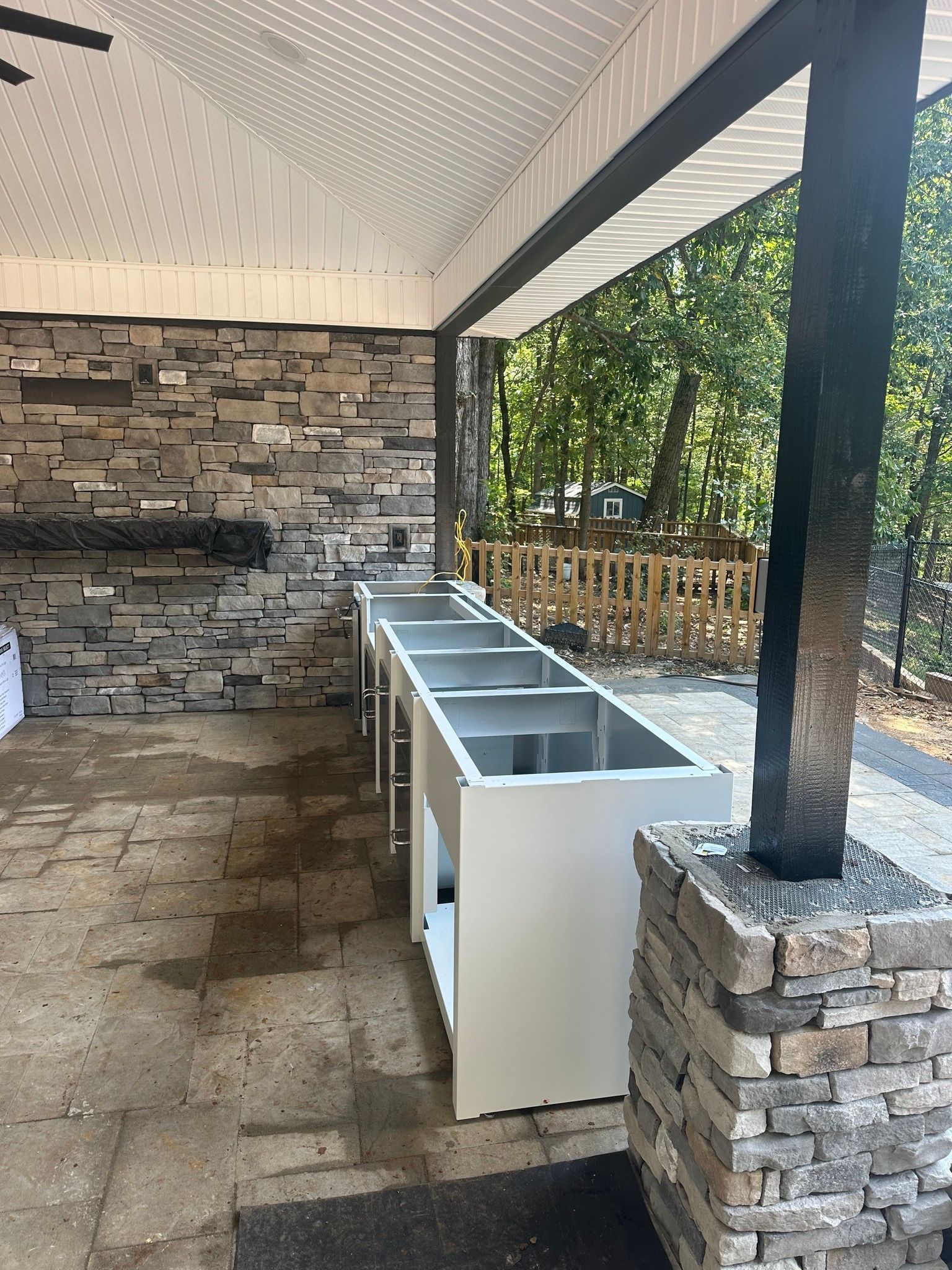 Covered outdoor sink station with five white basins, stone walls, and a black support post overlooking trees.