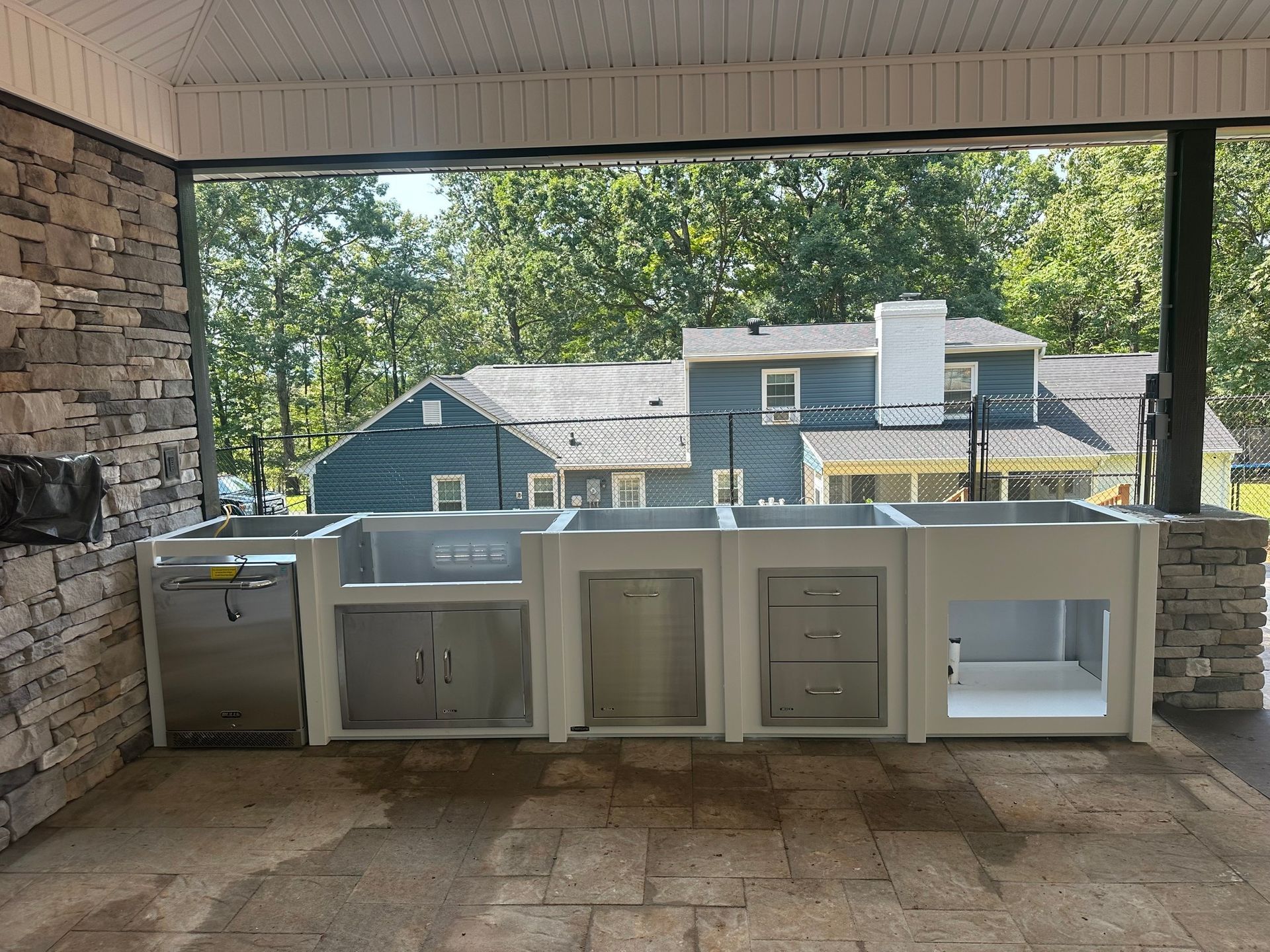 Outdoor covered patio with a row of outdoor kitchen cabinets and a sink against a stone wall.