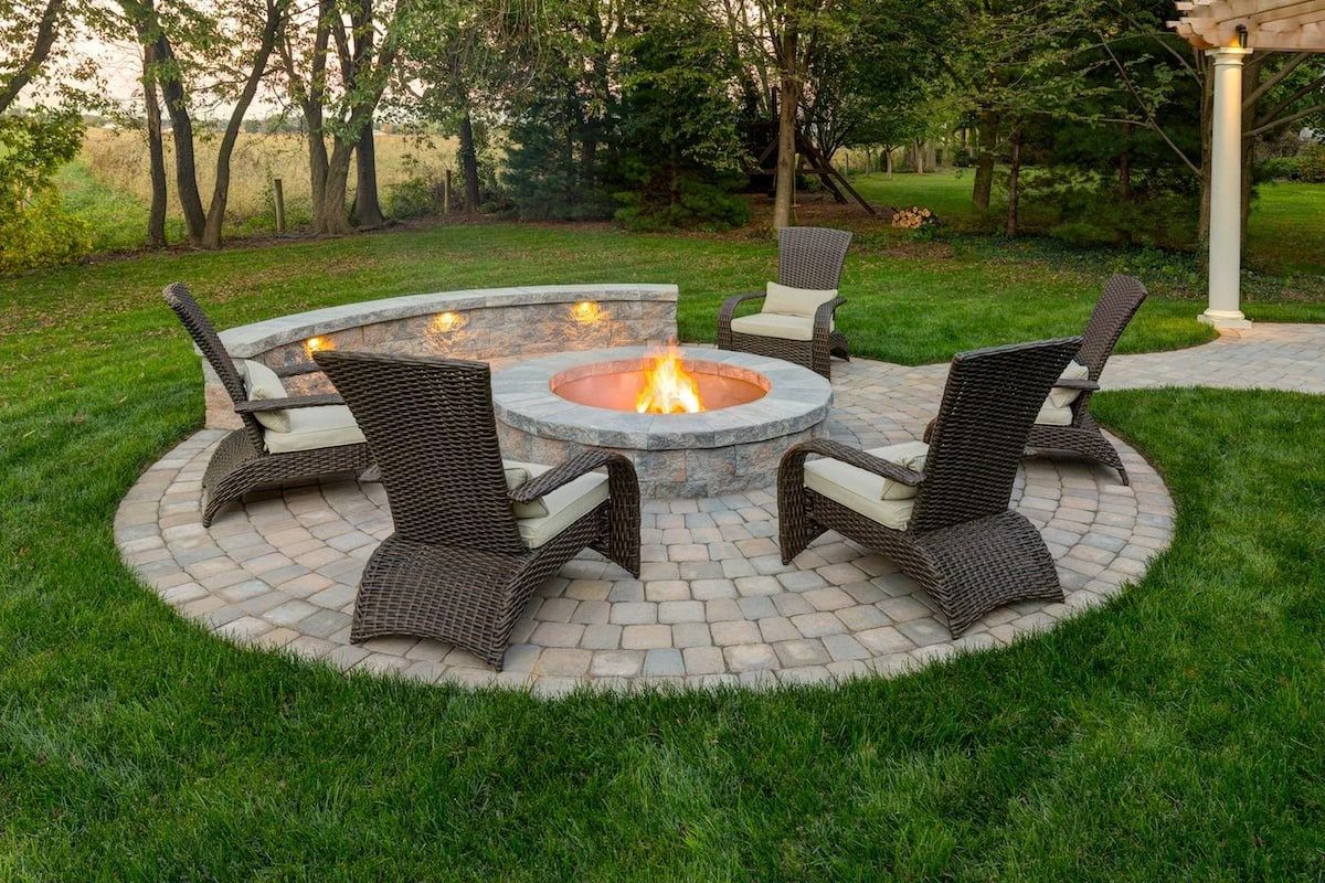 Fire pit with seating in a grassy backyard, surrounded by a paved patio and stone wall.