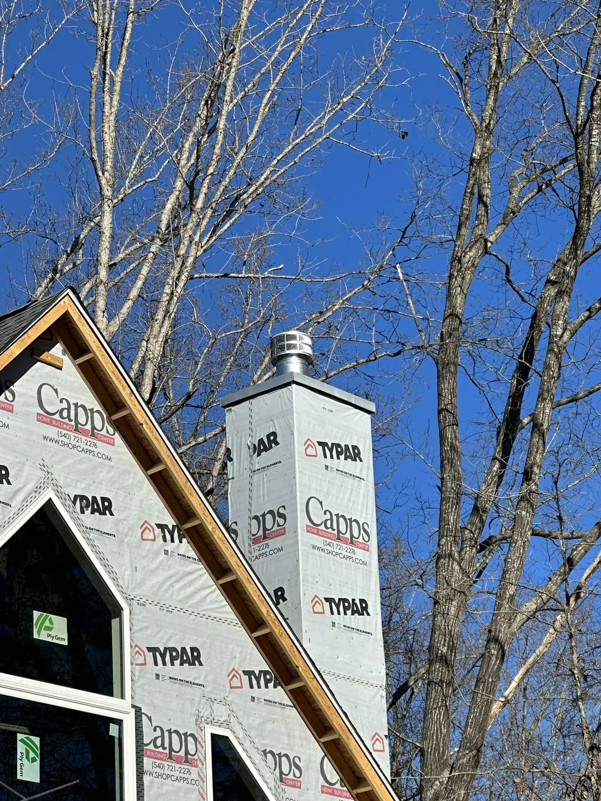 New house under construction, with Typar wrap, chimney, and bare tree branches against a blue sky.