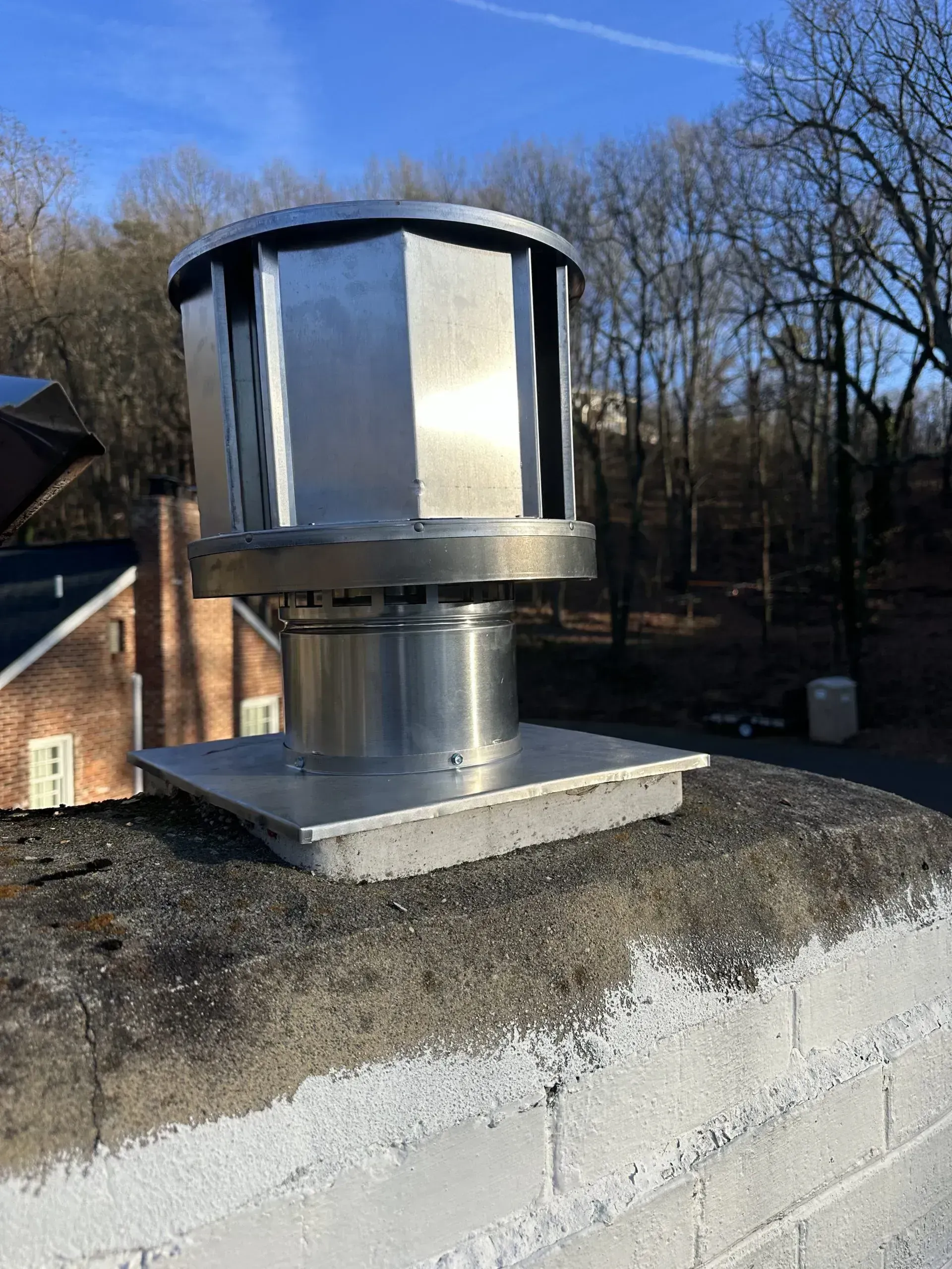 Silver rooftop vent on a brick chimney against a backdrop of trees and a blue sky.