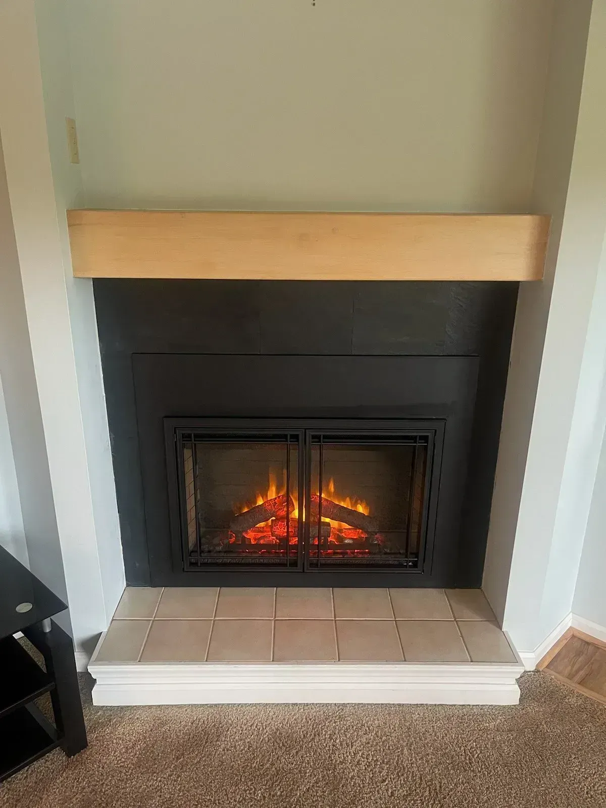 Electric fireplace with black surround, wooden mantel, and tan tile hearth.