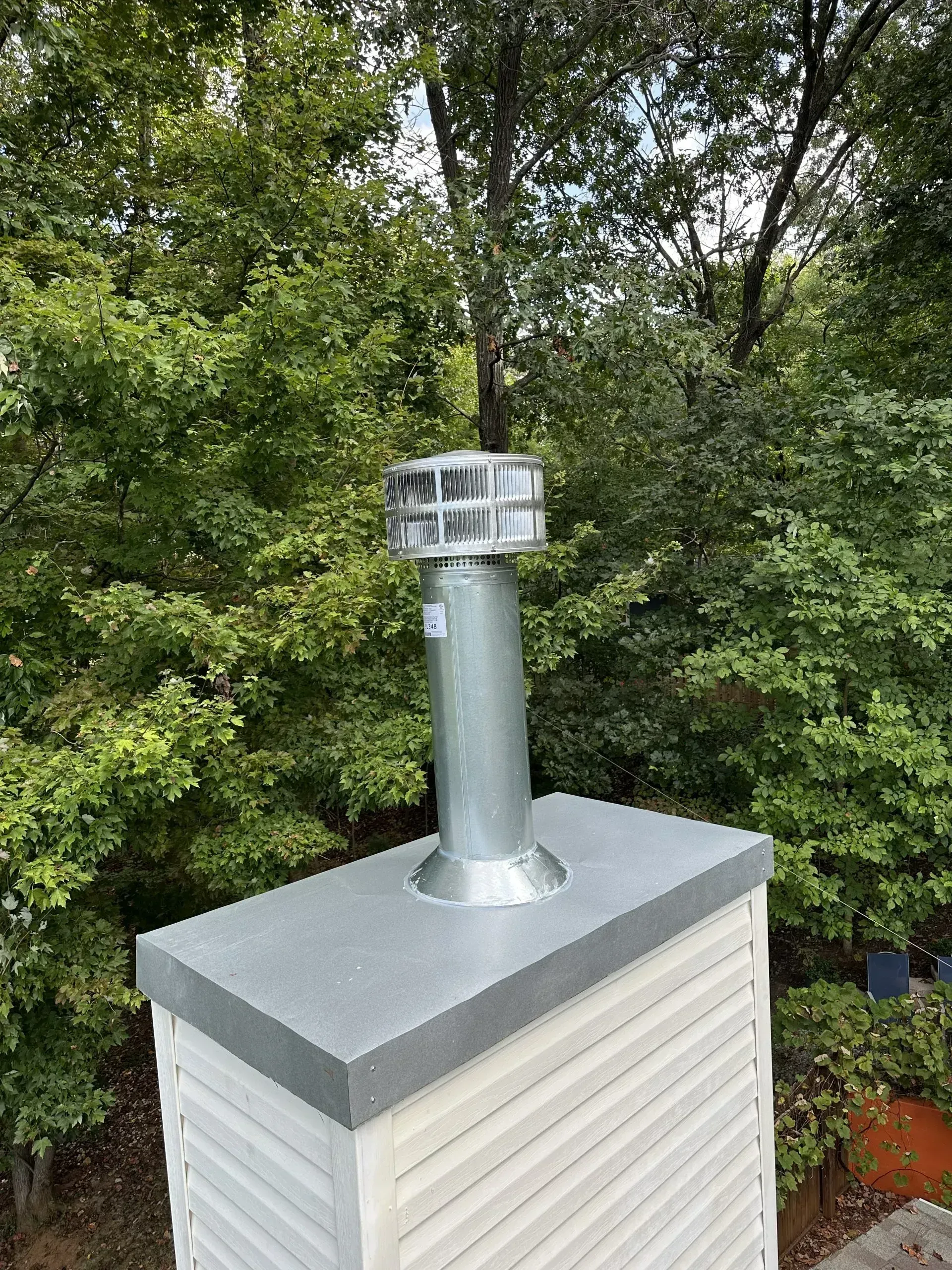 Metal chimney cap and pipe on a white-sided structure against a backdrop of green trees.