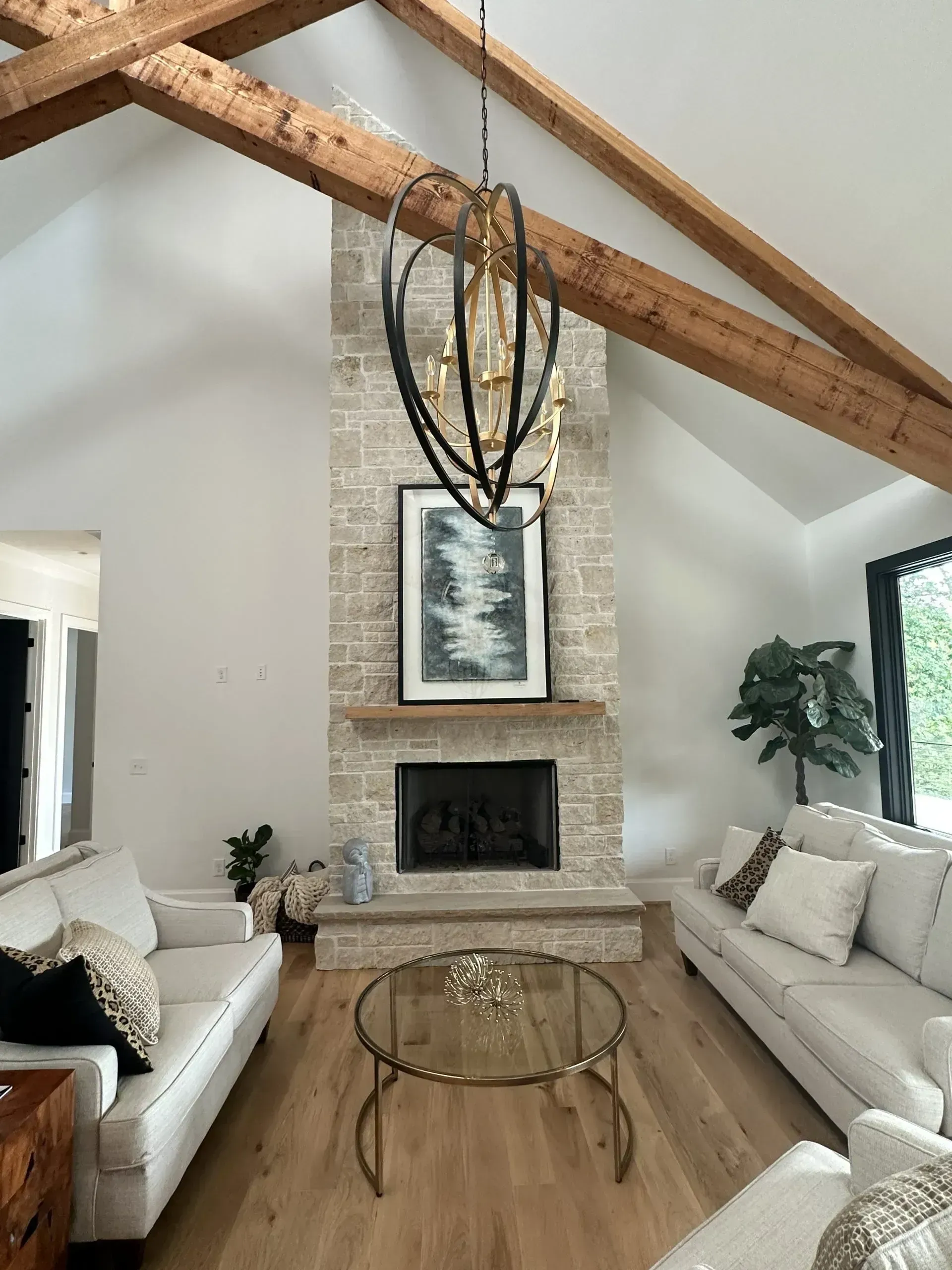 Living room with stone fireplace, wood beams, and couches. Black chandelier hangs above.