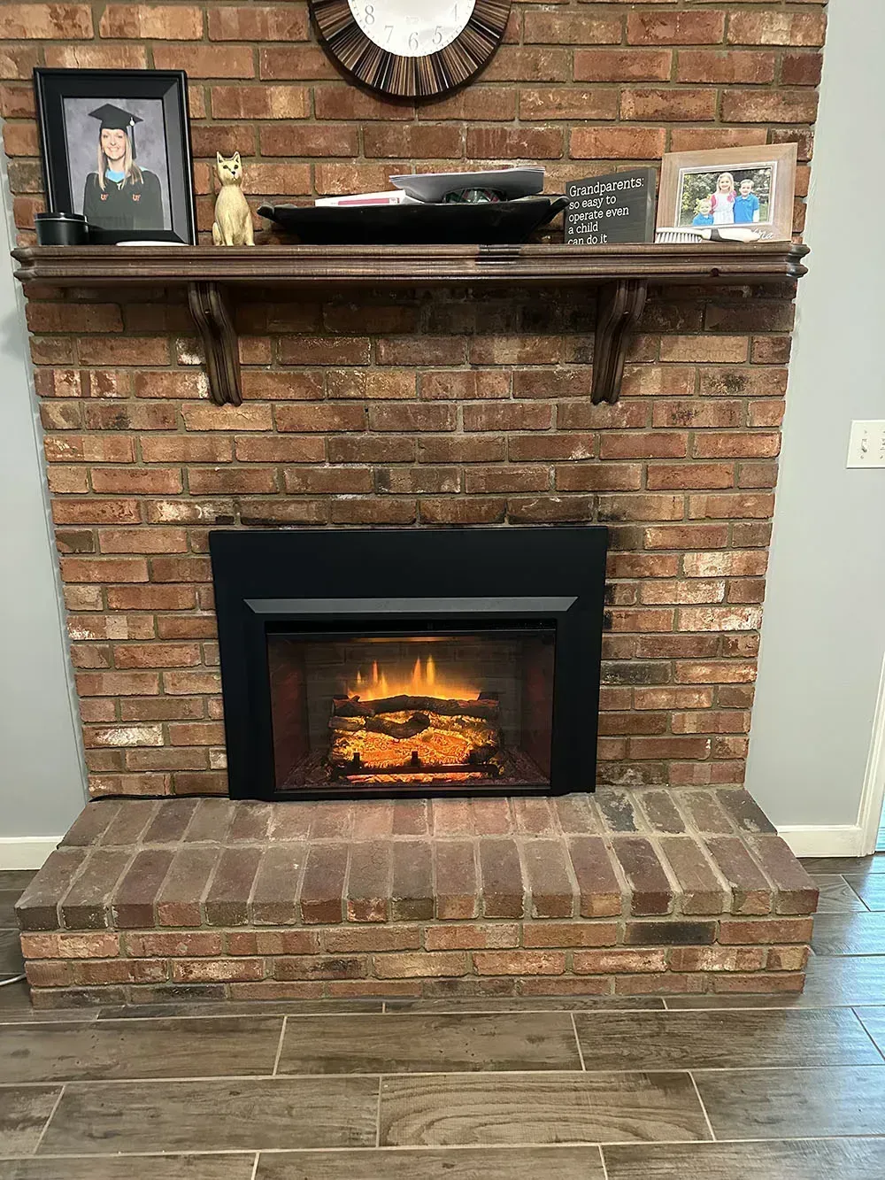 Brick fireplace with mantel holding decor: picture frame, bowl, and mirror. Electric fireplace insert has glowing embers.