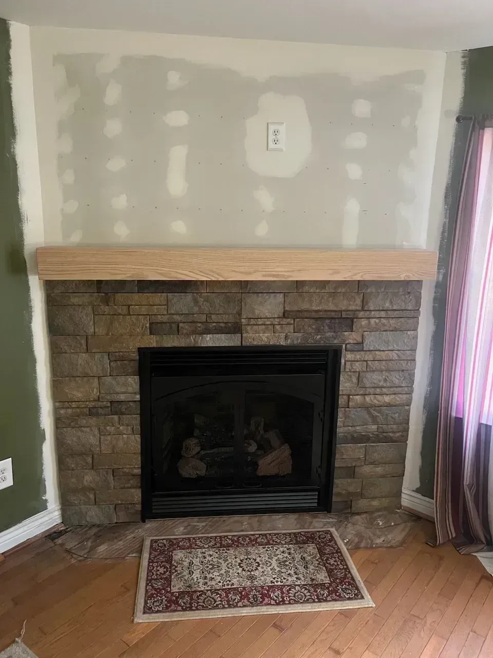 Fireplace with stone facade, wooden mantel, and rug. Drywall surrounds are unfinished.