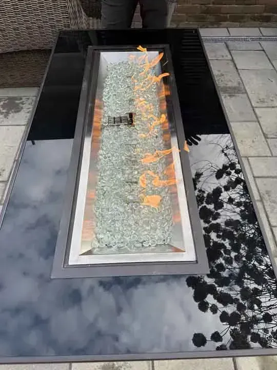 A rectangular fire pit table with flames rising above light green fire glass, on a tiled patio.