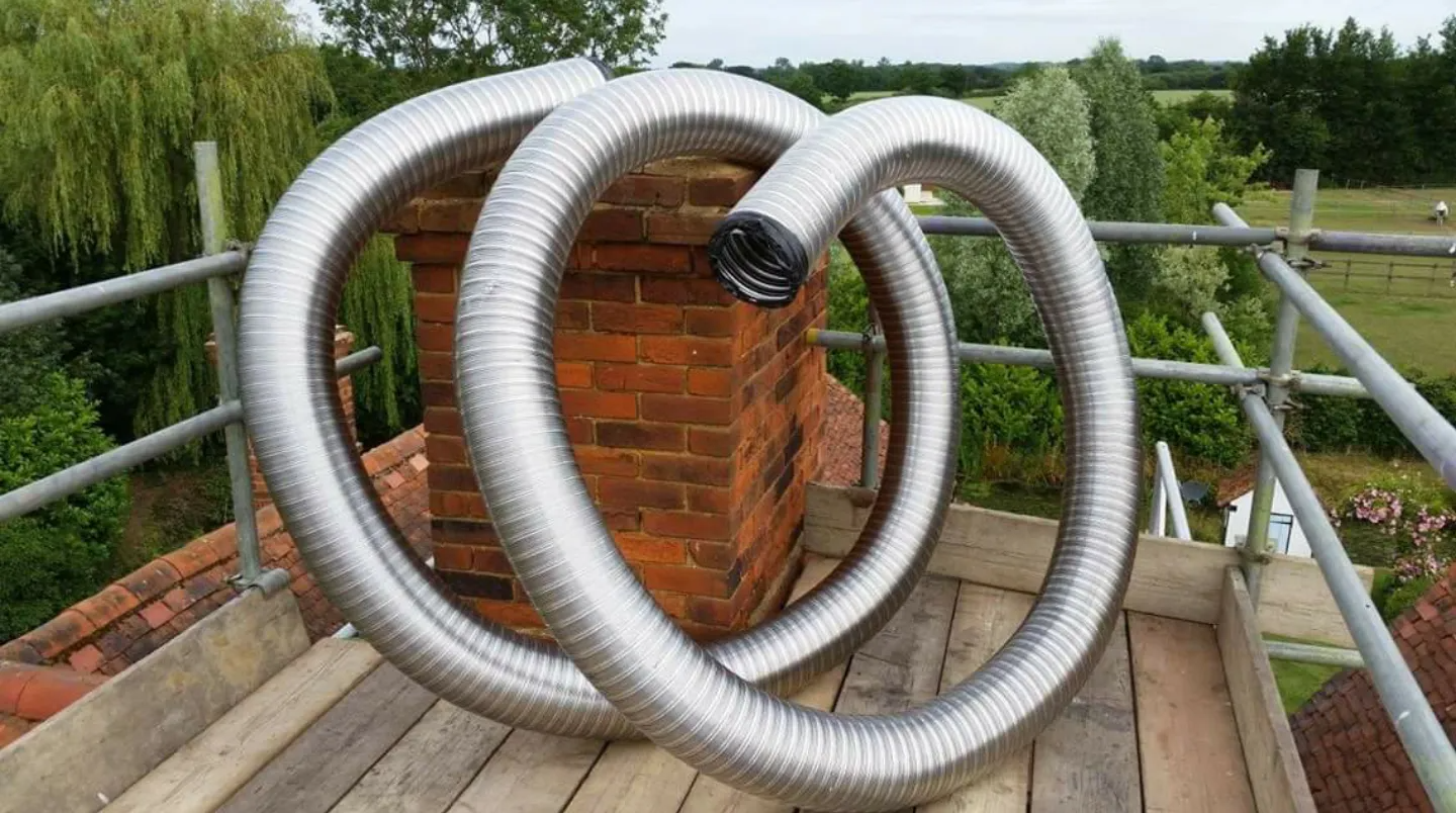 Coiled metal chimney liner on a rooftop, next to a brick chimney and scaffolding, with a green landscape in the background.