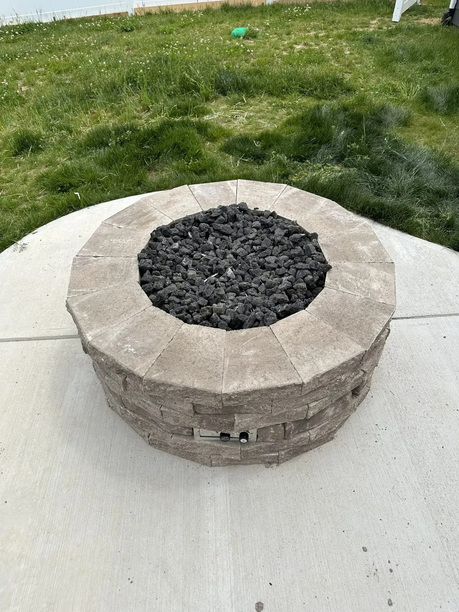 Stone fire pit with black rocks in the center, on a concrete patio, with green grass in the background.