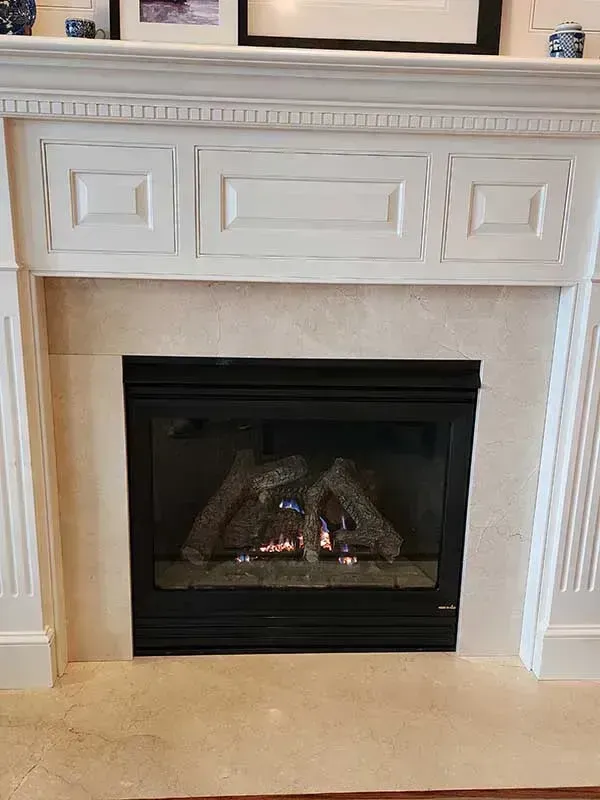 Fireplace with lit gas flames, black frame, light marble surround, white mantle.