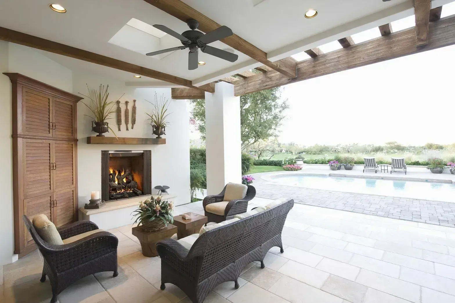 Patio with fireplace, seating, ceiling fan, and pool view. Beige tile floor, wooden beams, sunny outdoor setting.