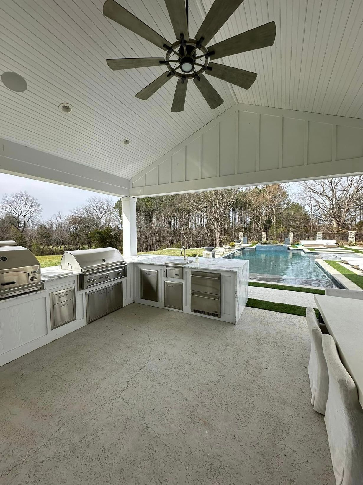Outdoor kitchen under a white-paneled ceiling with a fan, facing a pool through the opening.