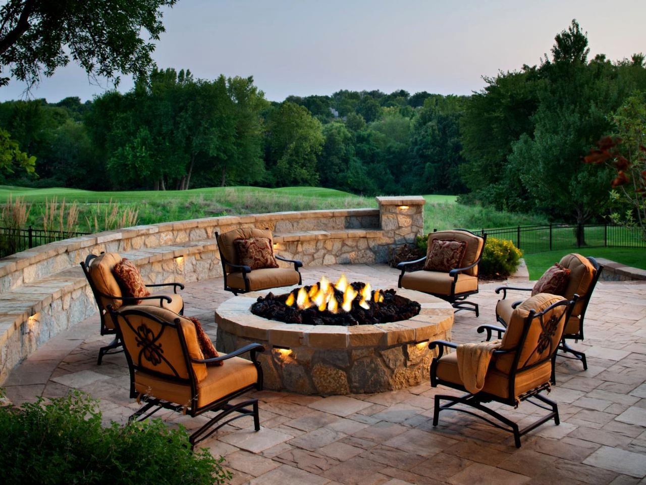 Stone fire pit surrounded by chairs on a patio, overlooking a green landscape.