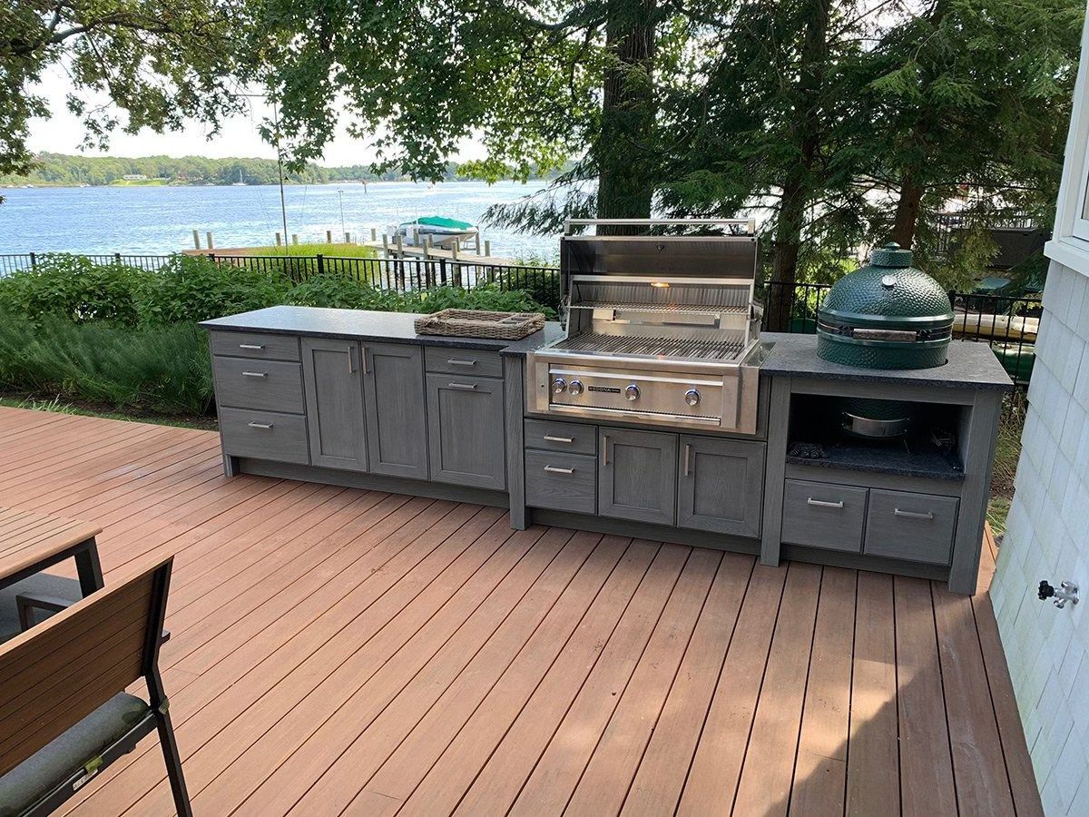 Outdoor grilling station on a wooden deck overlooking a lake with gray cabinetry and a stainless steel grill.