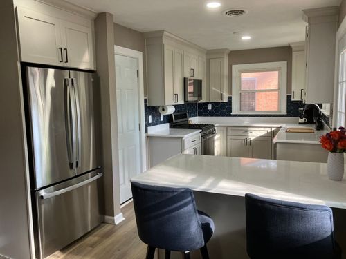 Bright white modern kitchen with stainless fridge, white island, and two dark stools by the window.