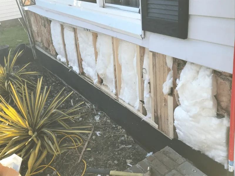 Damaged house siding with exposed insulation beneath a white window and black trim