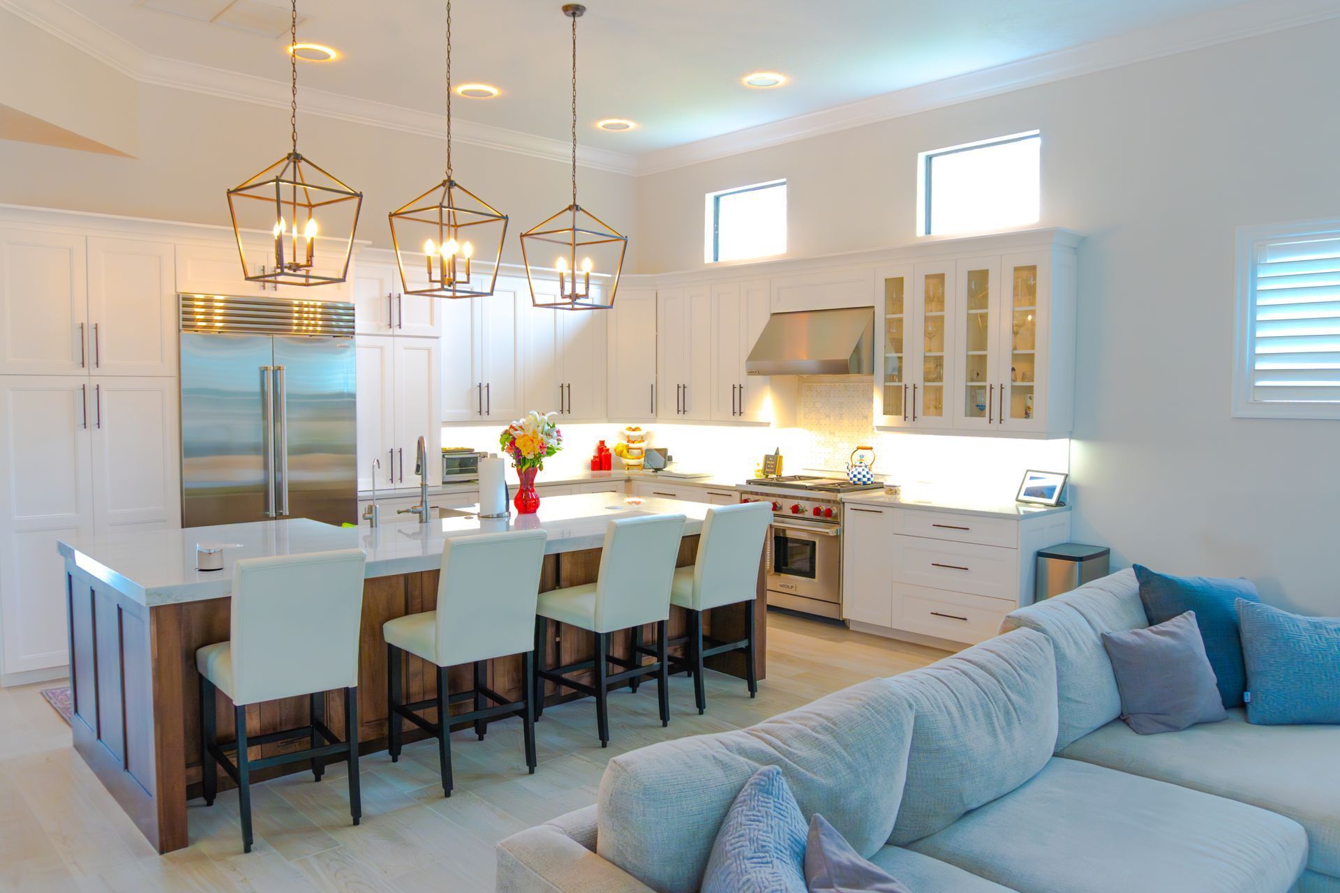 Modern kitchen with island, pendant lights, and stainless steel appliances. White cabinets, wooden accents, and a light-colored sectional sofa.