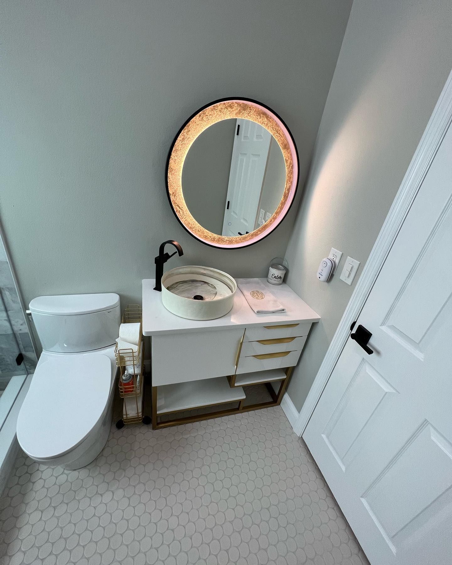 Modern bathroom with white vanity, round mirror, and toilet; gray walls and hexagonal tiles.