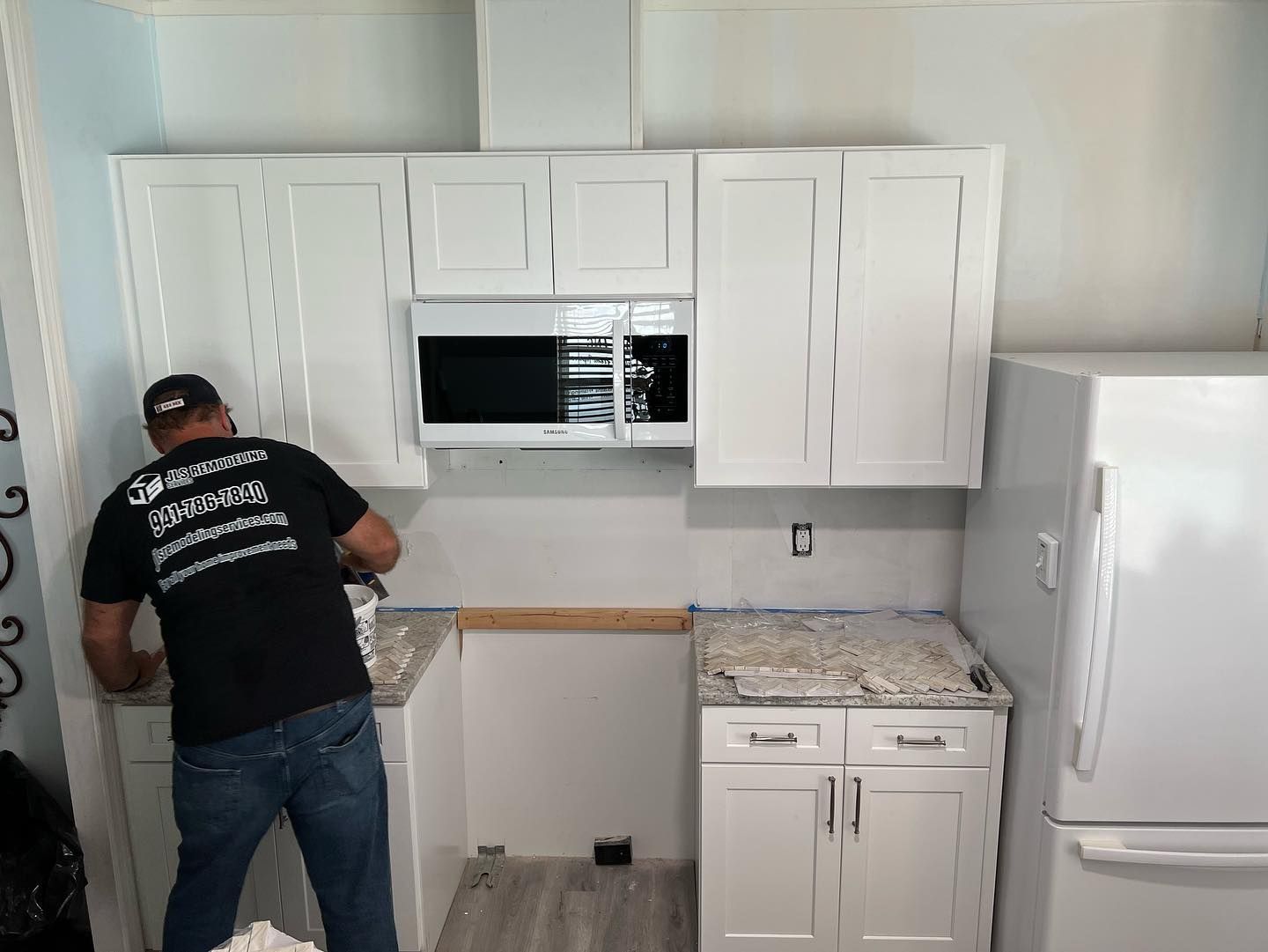 Man installing a kitchen backsplash with white cabinets and microwave. White refrigerator is on the right.