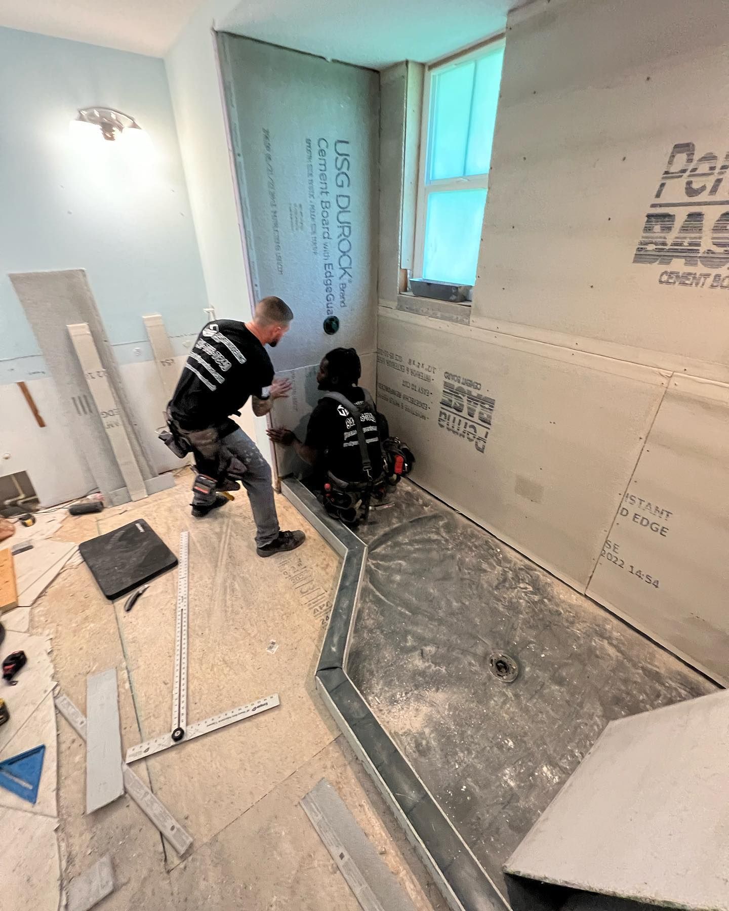 Two workers installing shower wall panels in a bathroom remodel; one uses a circular saw.