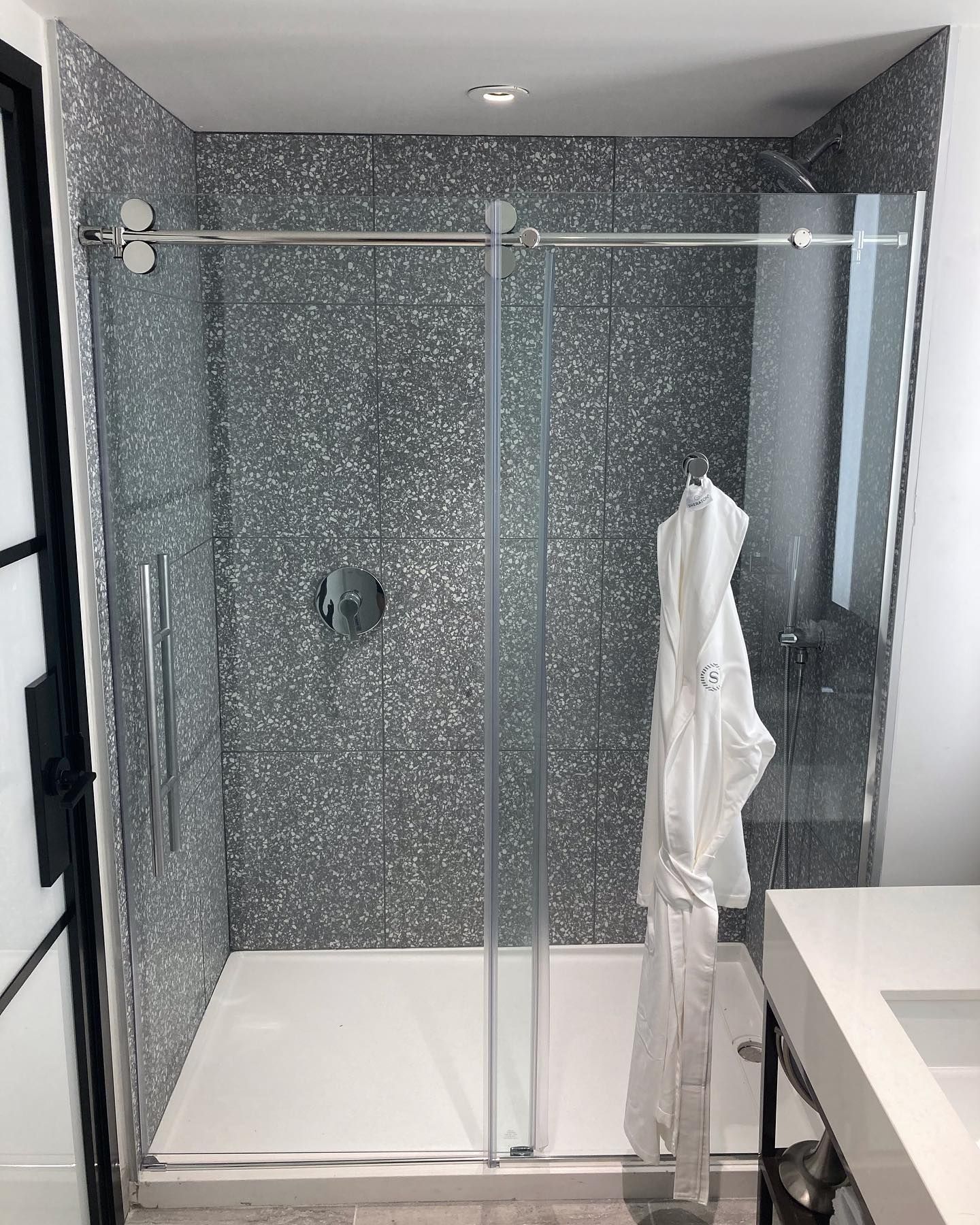 Modern shower stall with gray speckled tile, glass doors, and a white robe.
